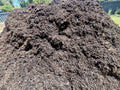A large pile of dark brown compost or mulch outdoors, with some greenery and fencing visible in the background.