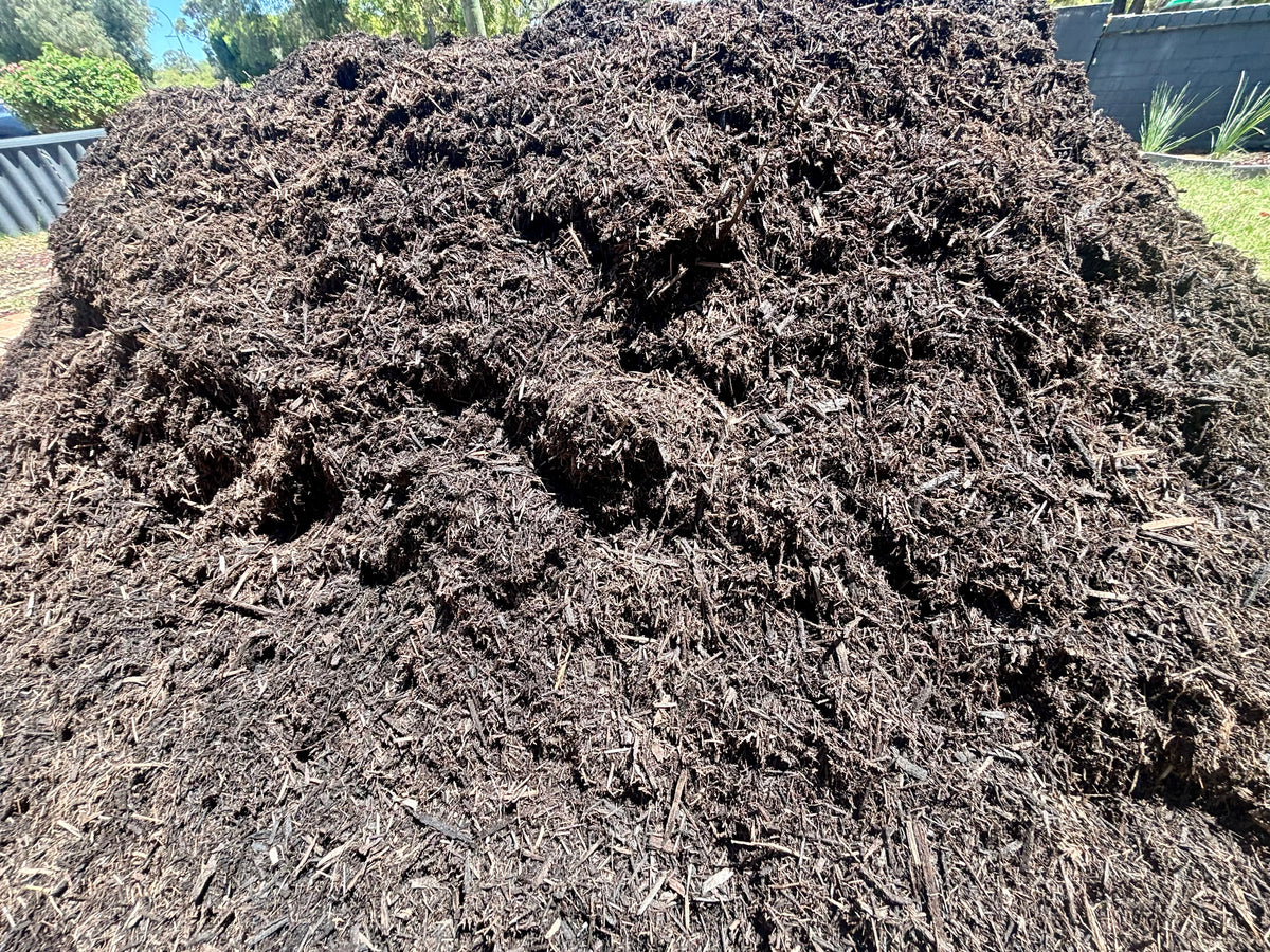 A large pile of dark brown compost or mulch outdoors, with some greenery and fencing visible in the background.